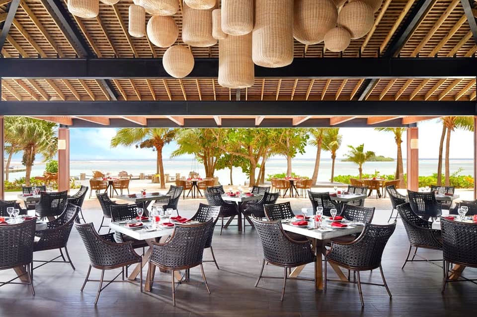 Beachfront dining pavilion with woven pendant lights, palm trees, ocean view, and round tables