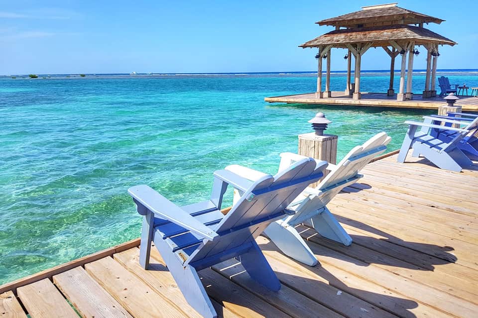 Wooden dock with white and blue Adirondack chairs facing crystal-clear turquoise water and gazebo