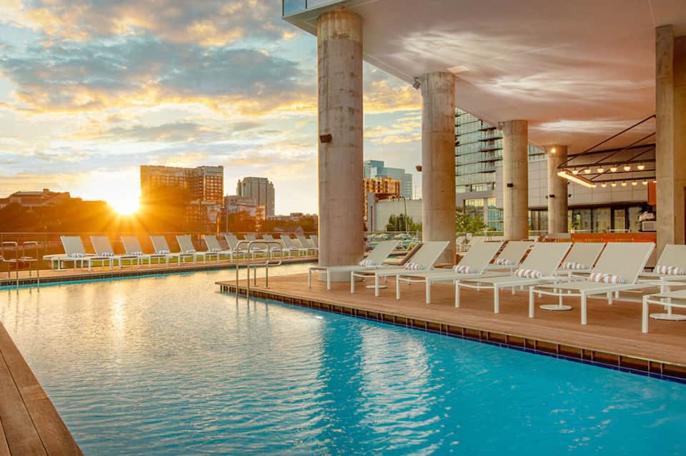 Indoor pool with natural light, white lounge chairs, and sunset city skyline views through large windows