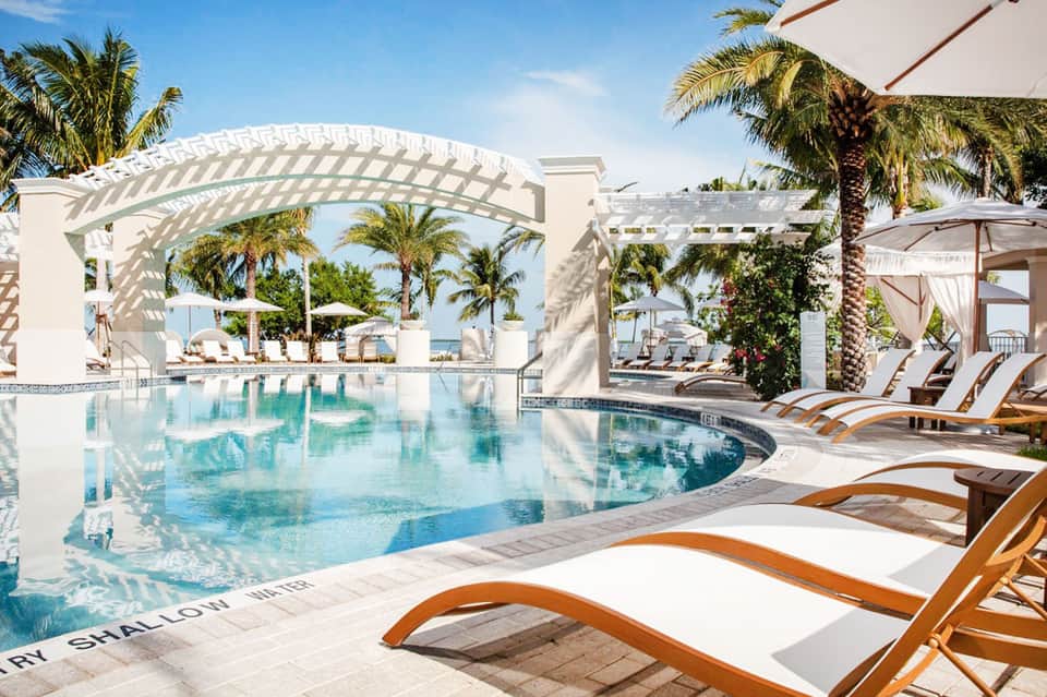 Luxury resort pool with white pergola, palm trees, lounge chairs, and ocean view on sunny day