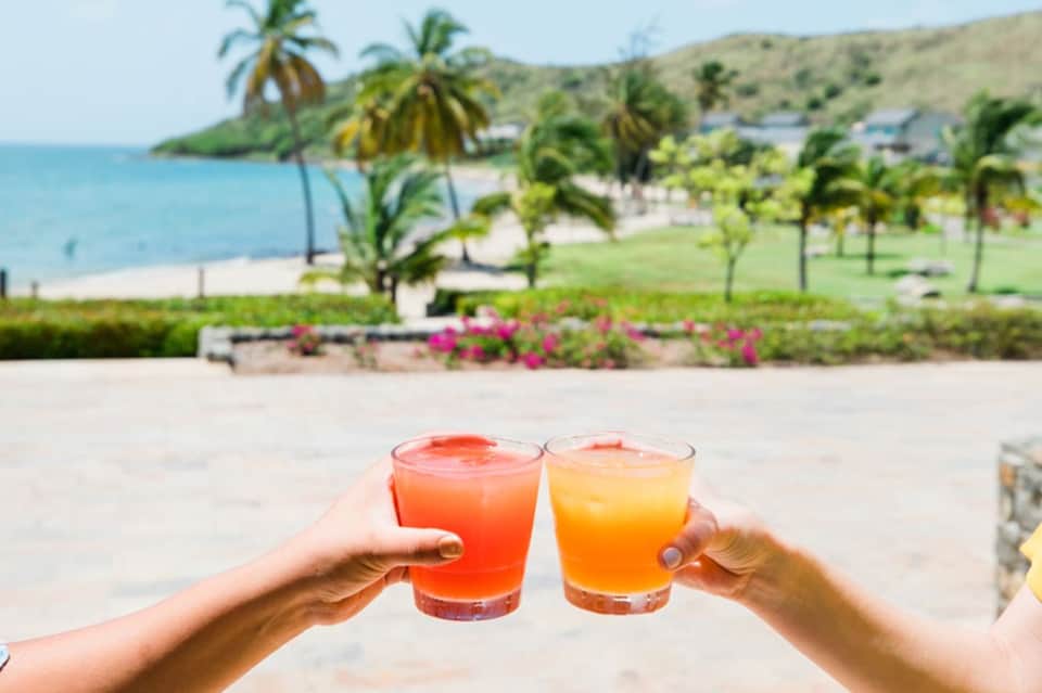 Two colorful drinks held up on beach with ocean, palm trees, and coastline in background