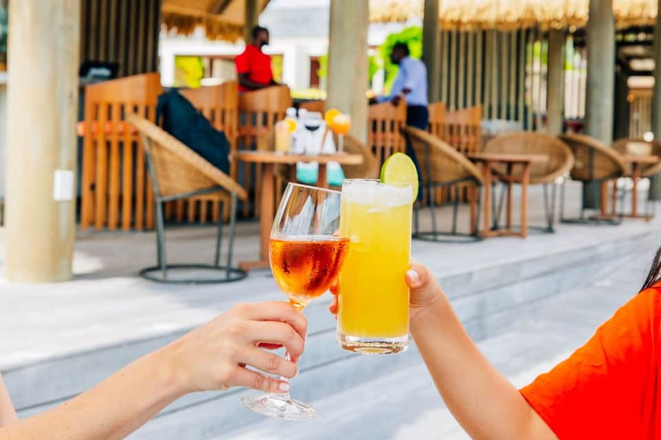Two colorful cocktails held outdoors on a resort terrace with wooden chairs and tropical setting