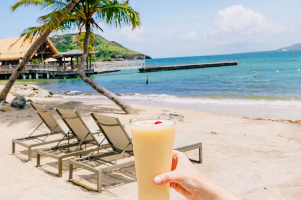Hand holding tropical cocktail on beach with palm trees and tiki hut