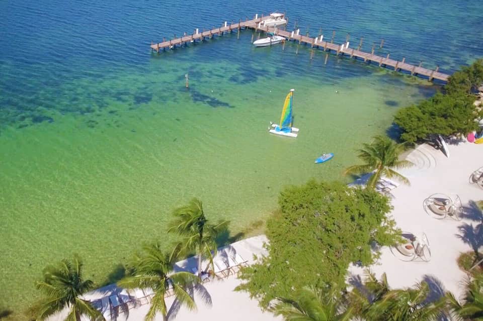 Aerial view of tropical beach with clear turquoise water, wooden dock, sailboats, and palm trees