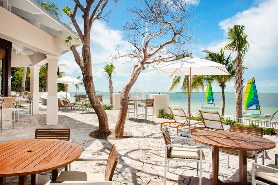 Beachfront resort terrace with wooden tables, white umbrellas, and sailboats on turquoise water