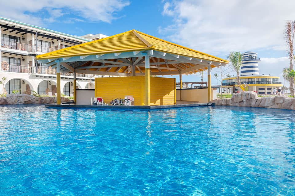Yellow and teal poolside cabana structure over bright blue resort pool with palm trees and waterfront buildings