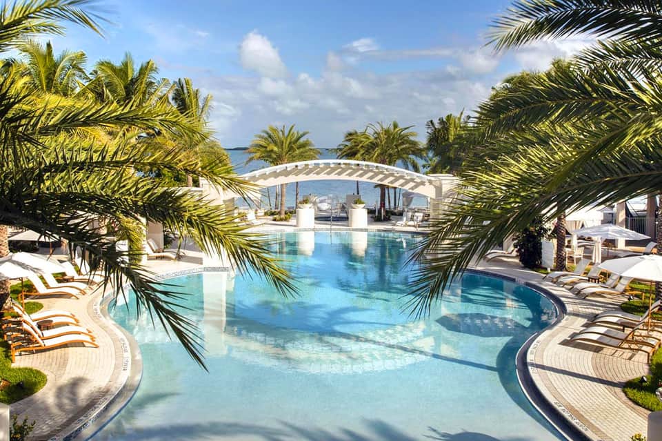 Luxurious resort pool with curved pergola, palm trees, loungers, and ocean view in background