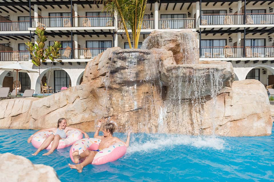 Children on inflatable pool floats near large rock waterfall feature in resort pool