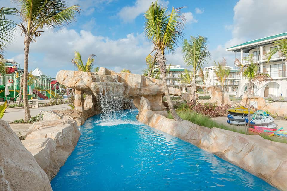 Resort waterpark with bright blue lazy river flowing through stone archway with palm trees and hotel buildings