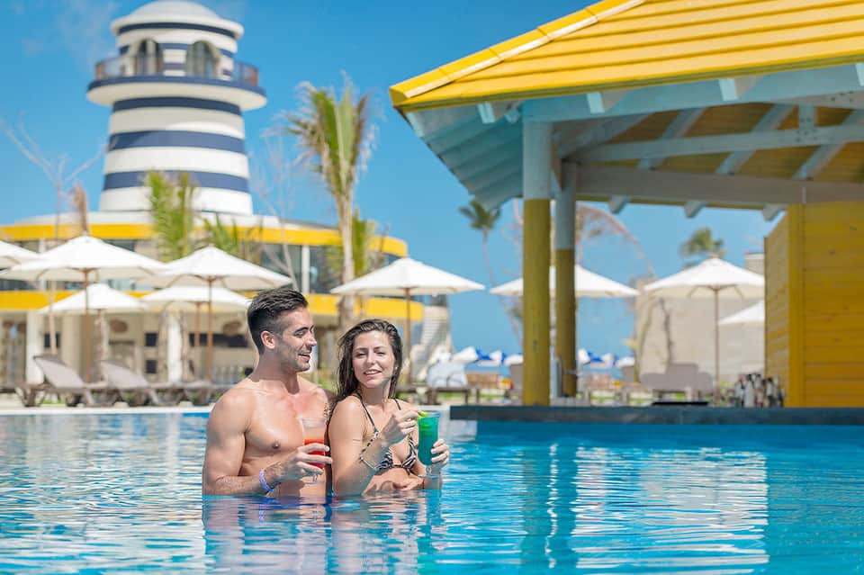 Couple enjoying drinks in resort pool with striped umbrella and lighthouse-style building