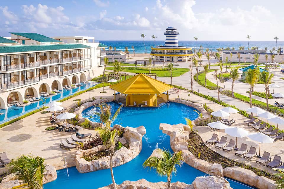 Beachfront resort with blue lazy river, yellow roof structure, palm trees, and ocean view