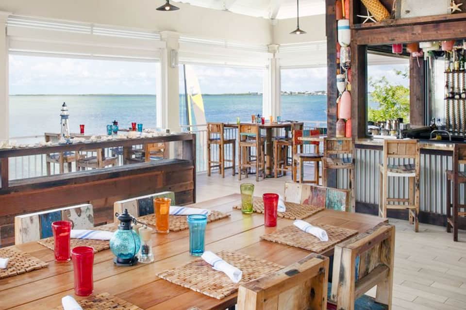 Waterfront dining room with large windows overlooking ocean, wooden tables with red candles, and nautical decor.