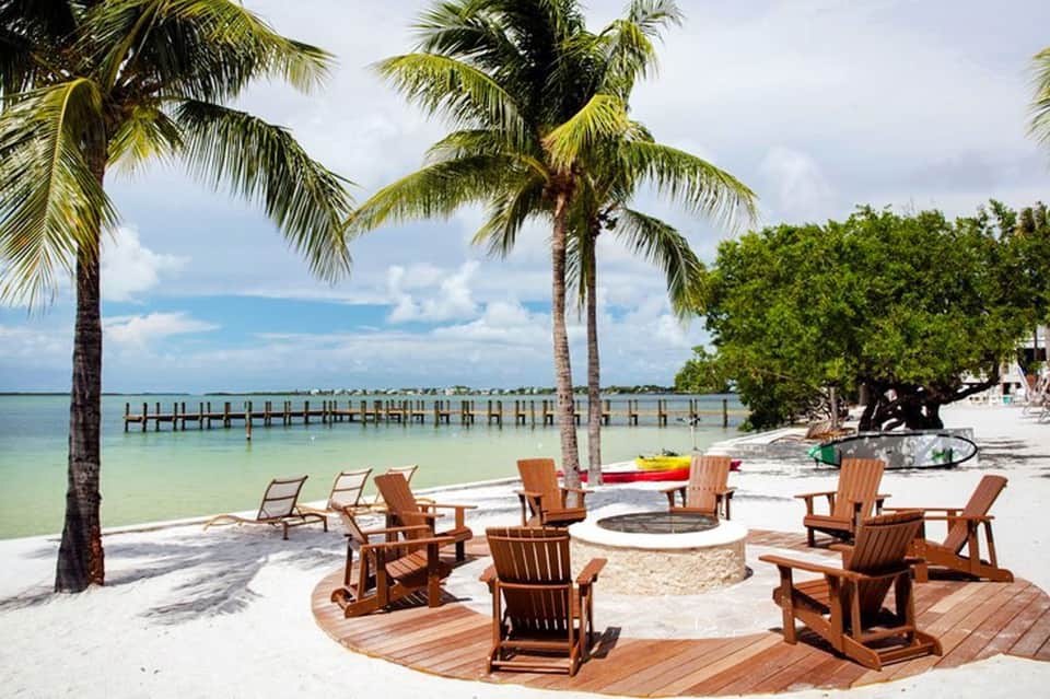 Beach resort with wooden Adirondack chairs around fire pit, palm trees, and wooden pier extending into turquoise water