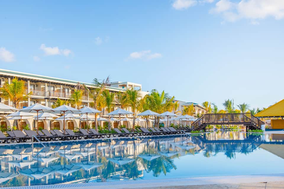 Resort pool with loungers under umbrellas, palm trees, and beachfront building