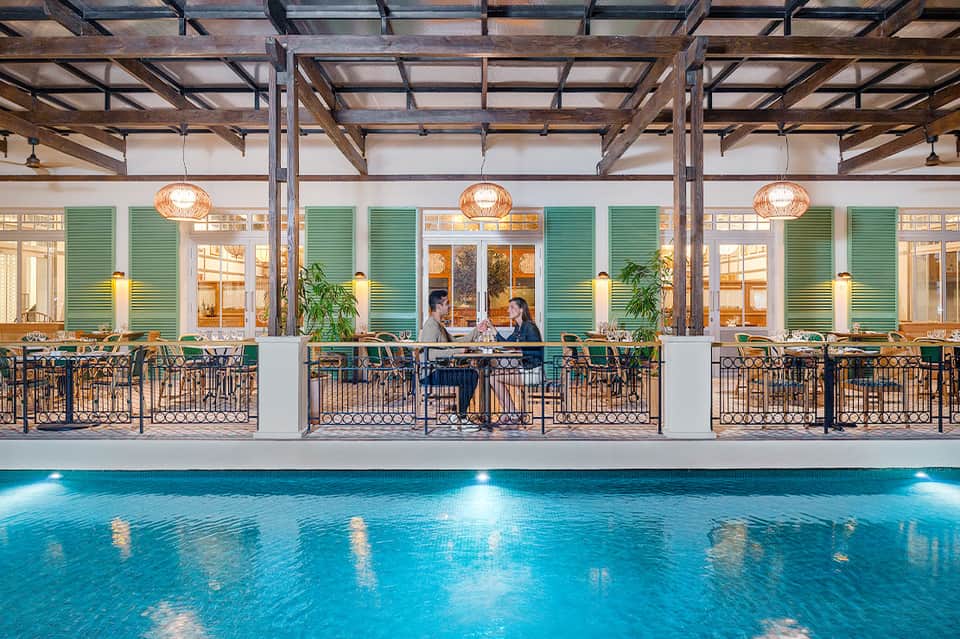 Indoor pool with green shuttered windows, hanging lights, and couple dining poolside