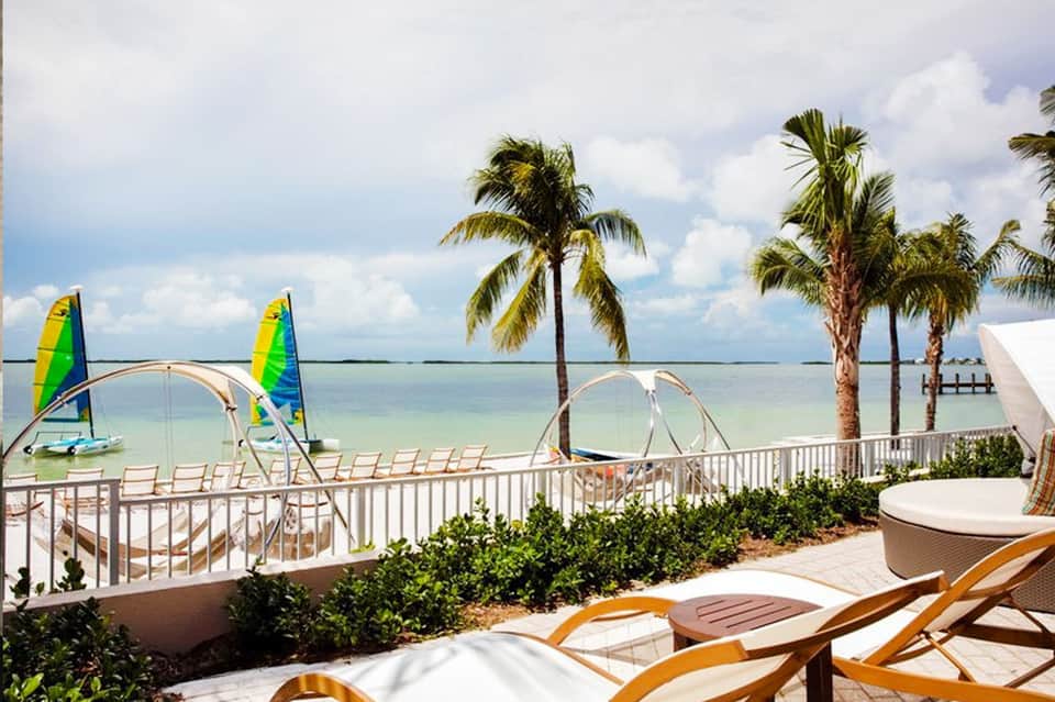 Beachfront resort deck with lounge chairs, sailboats, palm trees, and ocean view