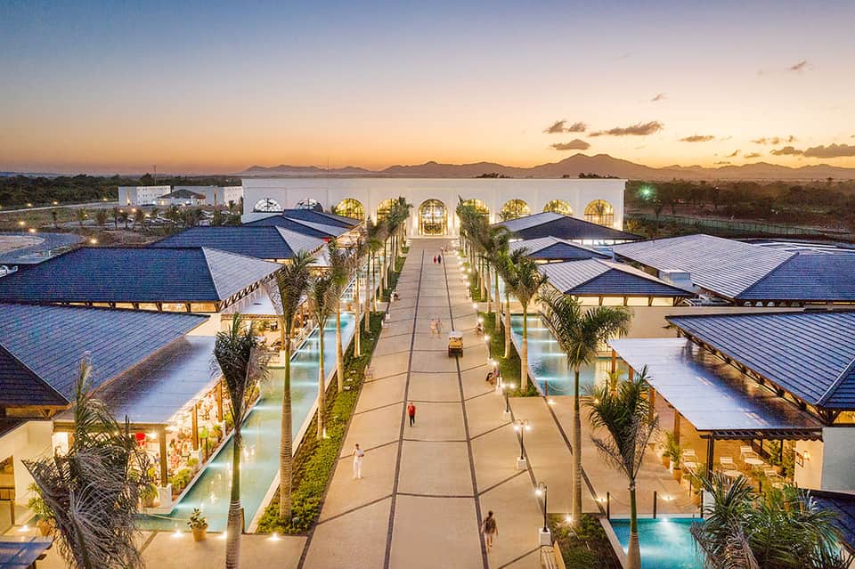 Aerial view of luxurious resort complex with blue-roofed buildings, palm trees, and central promenade at sunset