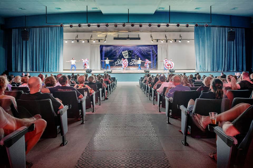 Theater auditorium with red seats, blue curtains, and stage performance with dancers and neon signage