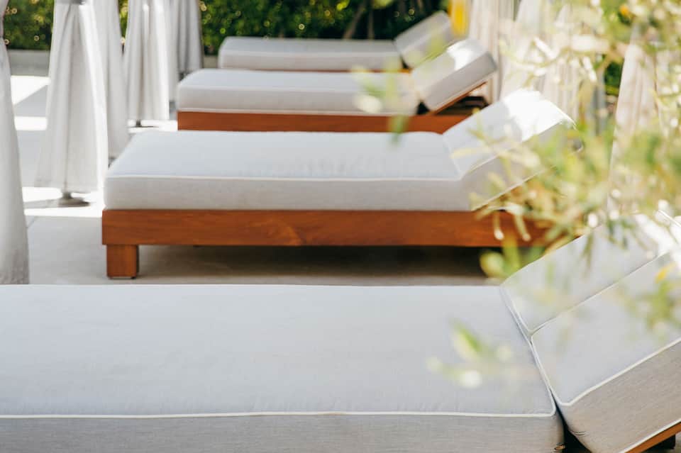 Row of wooden loungers with white cushions lined along poolside, surrounded by greenery