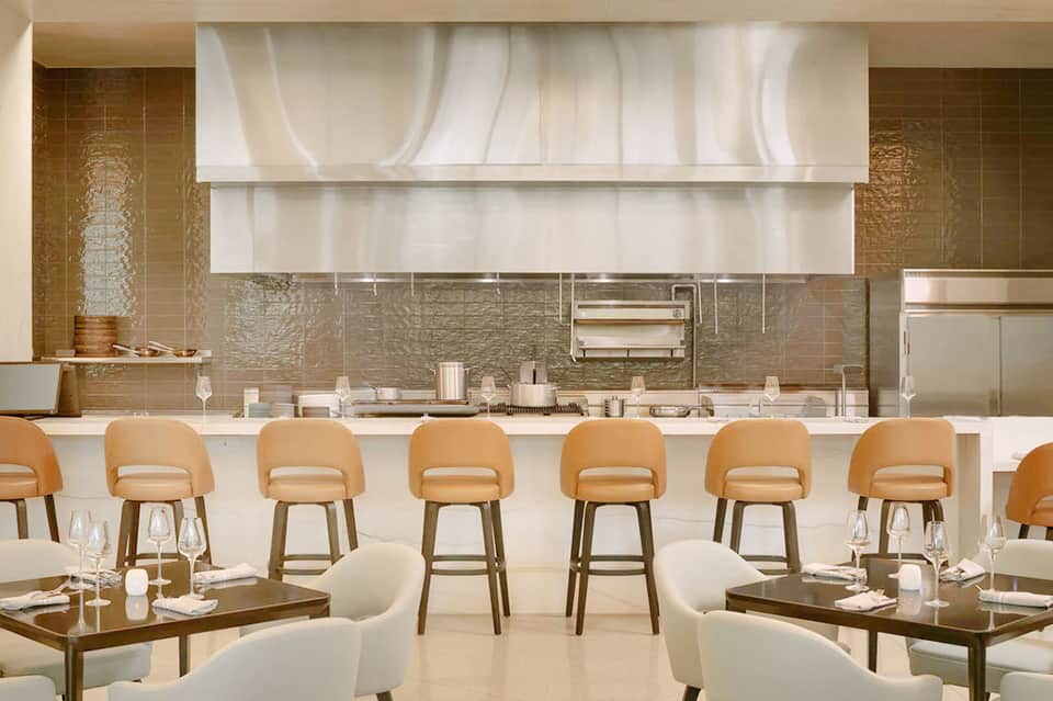 Modern restaurant kitchen and bar with white shelving, tan bar stools, and contemporary design