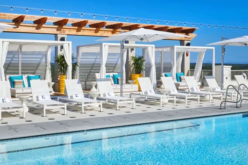 Resort pool with white loungers under wooden pergola with string lights, ocean view in background on sunny day