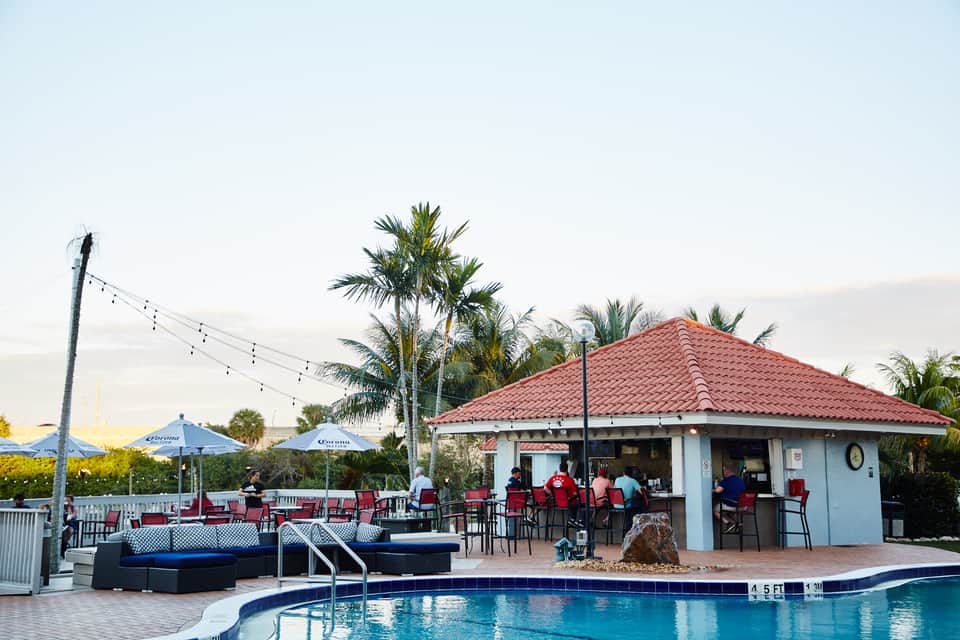 Poolside bar and lounge area with palm trees, red tile roof, and blue umbrellas