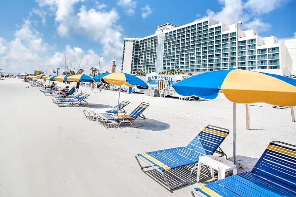 Sandy beach with blue and yellow umbrellas, lounge chairs, and a tall beachfront hotel building in the background