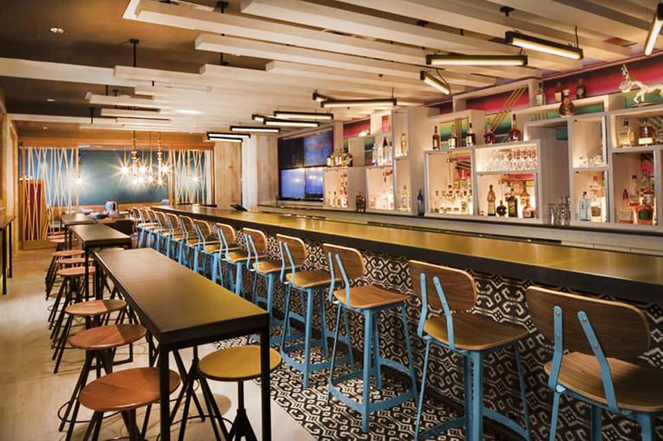 Modern bar interior with high seating, geometric patterns, pendant lighting, and shelved beverages