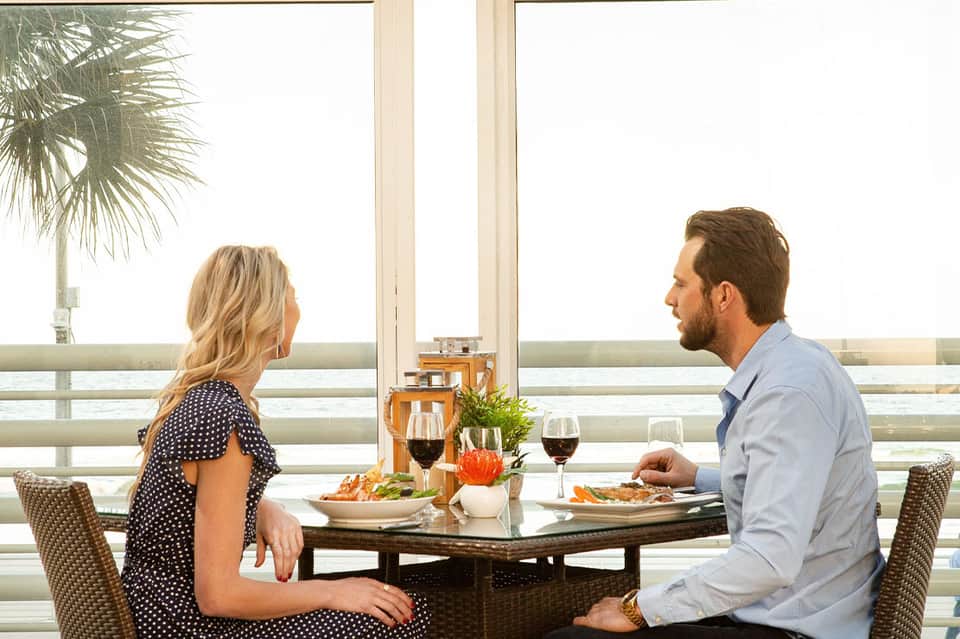 Couple dining on beachfront terrace with ocean view, wine glasses and appetizers on table, palm tree visible