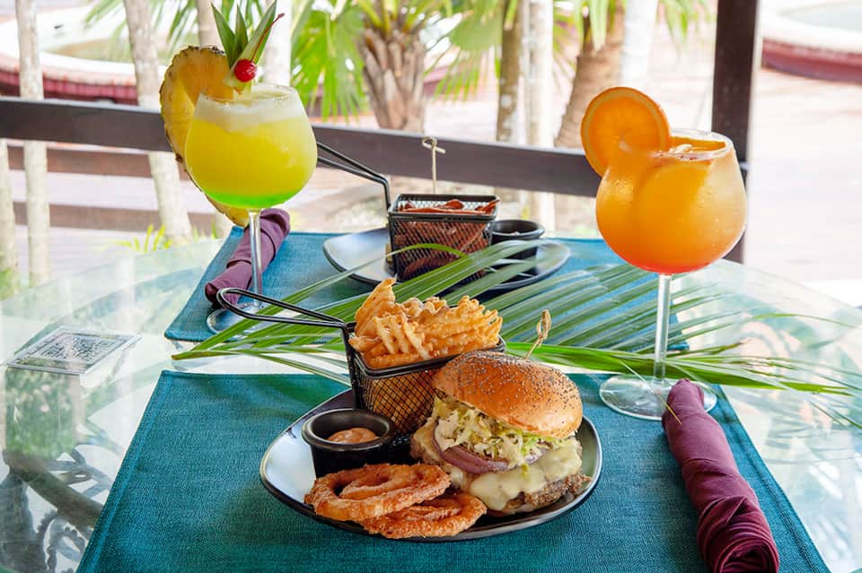 Tropical dining setup with colorful cocktails, burger and fries on blue-patterned placemats by palm trees