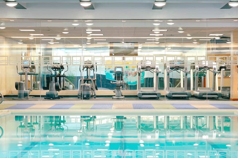 Indoor swimming pool with exercise equipment visible through glass partition and bright ceiling lights