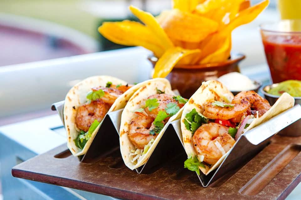 Three shrimp tacos with lime wedges and salsa on wooden serving board with fries