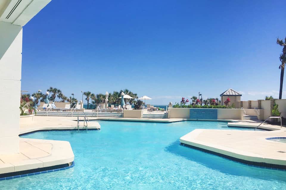 Outdoor resort pool with geometric design, palm trees, lounge areas, and ocean horizon view