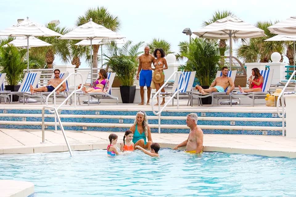 Resort pool area with families swimming, lounge chairs, umbrellas, and palm trees in background