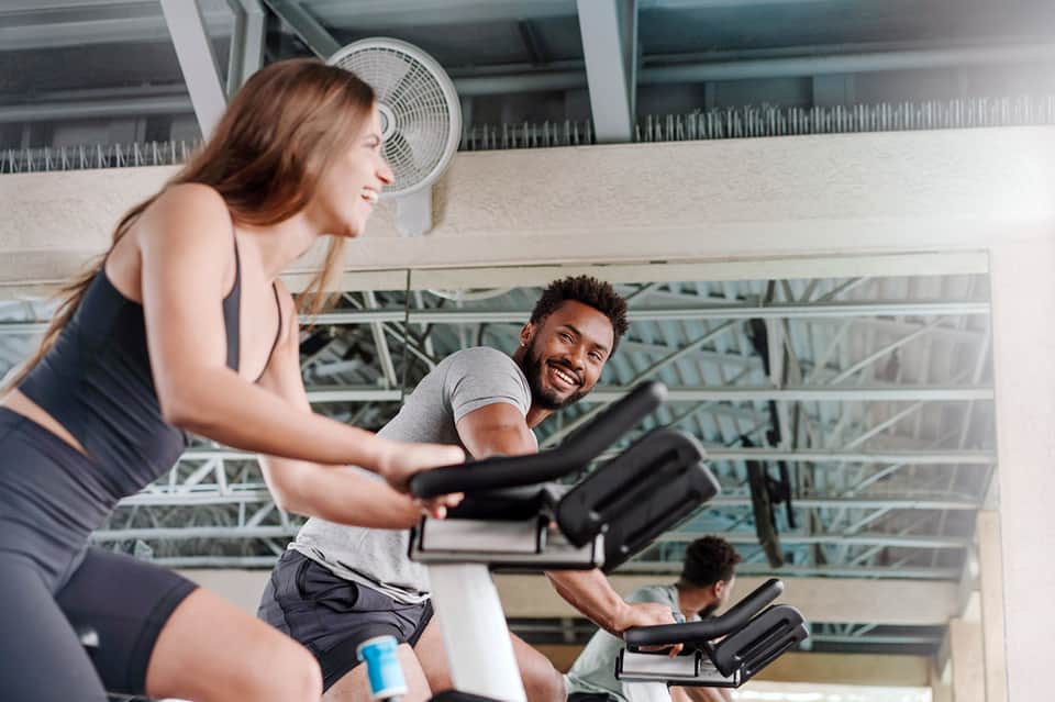 Fitness class with instructor and participants on stationary bikes in bright gym with exposed ceiling and industrial ventilation