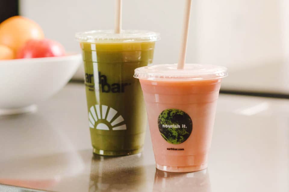 Two colorful beverages in clear cups with straws on a white counter with fruit bowl