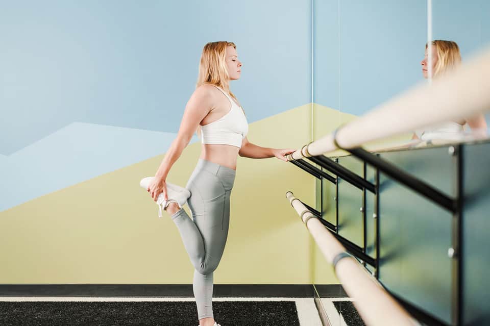 Woman in white sports bra stretching at ballet barre in modern gym studio