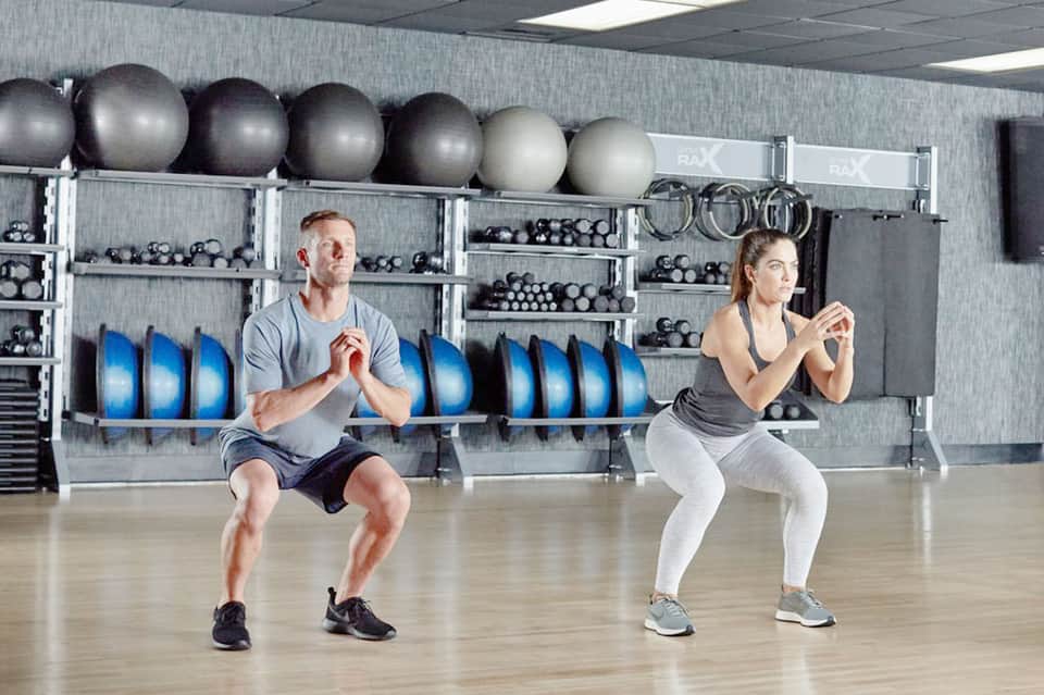 Couple performing squats in gym with exercise balls and dumbbells on shelving behind them