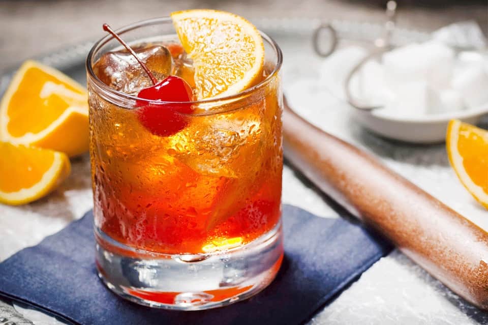 Cocktail with orange slice and cherry garnish on dark coaster with citrus slices nearby
