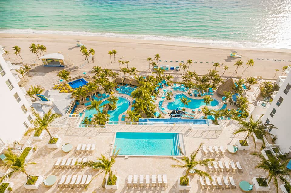 Aerial view of beachfront resort with multiple pools, palm trees, and loungers overlooking turquoise ocean