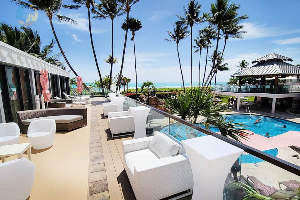 Beachfront resort pool deck with white lounge chairs, palm trees, ocean view, and modern architecture