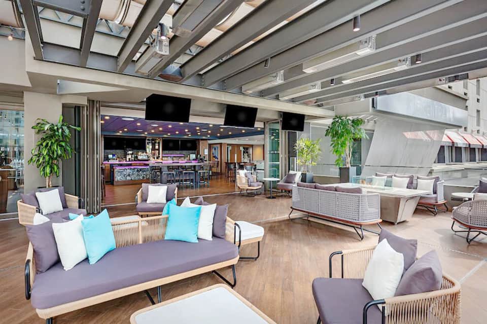 Modern resort lounge with purple seating, colorful pillows, wooden furniture, bar area, and indoor-outdoor layout with skylights