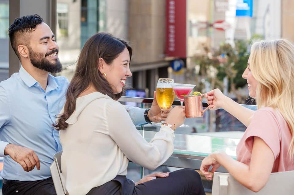 Three people toasting with colorful cocktails at an outdoor bar in an urban setting