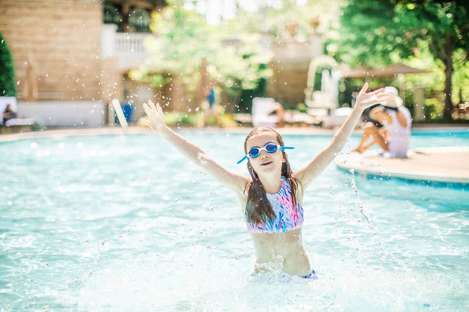 Child in pool wearing goggles with arms raised, splashing water in sunny resort setting
