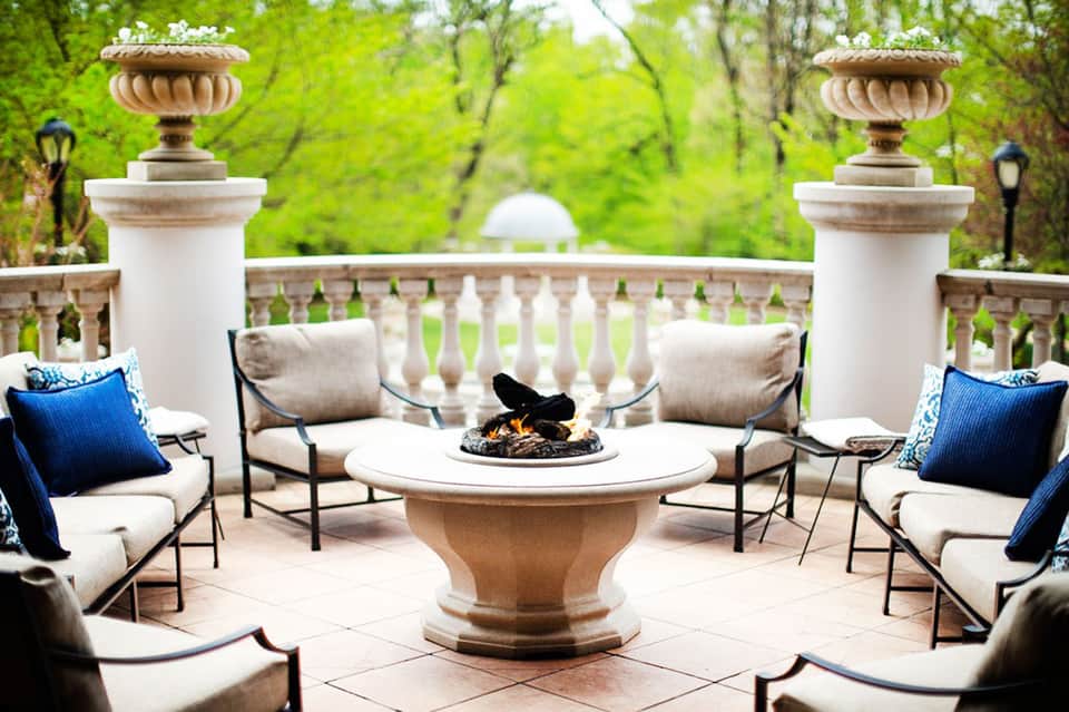Elegant outdoor patio with fireplace, cream seating, decorative urns, and manicured garden backdrop