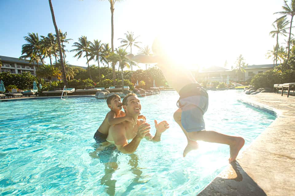 Family playing in resort swimming pool with palm trees and sun loungers in background