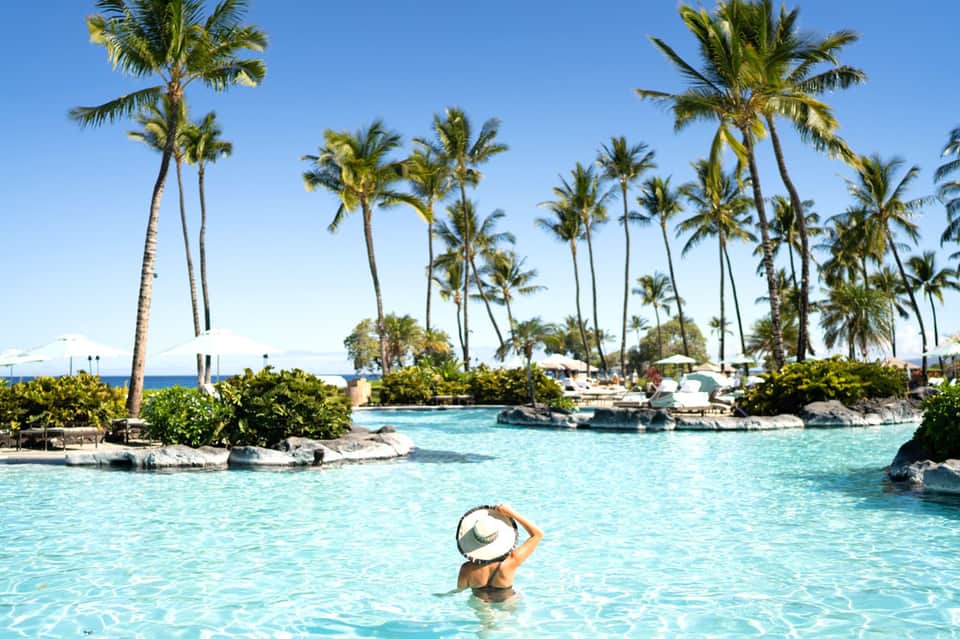 Person in pool wearing white hat, surrounded by tall palm trees and turquoise water