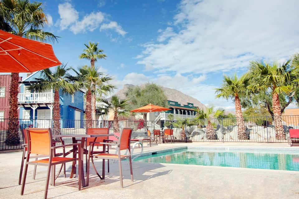 Resort pool area with palm trees, colorful umbrellas, lounge chairs, and desert mountains in background
