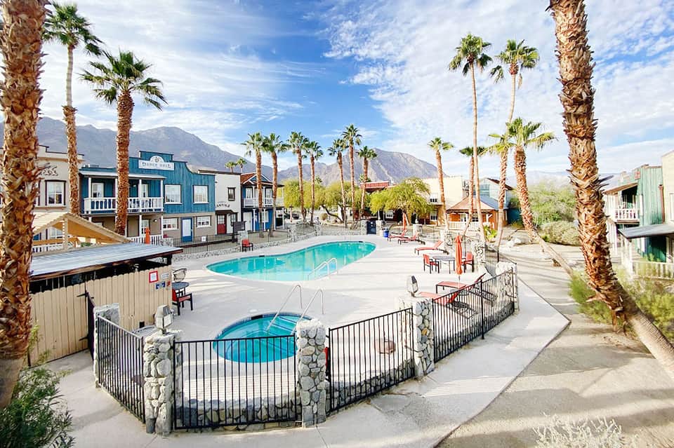 Desert resort pool area with palm trees, mountains, and colorful Western-style buildings