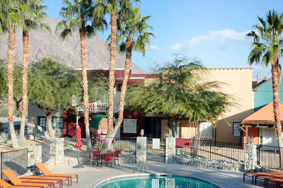 Desert resort courtyard with swimming pool, palm trees, colorful buildings, lounge chairs, and rocky mountain backdrop